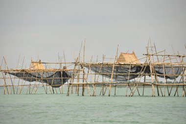 Bambu yapıları berrak suyun üzerinde yükselirler, bir kıyı bölgesinde balık kapanı görevi görürler. Güneş parıldıyor, gölgeler bırakıyor ve sakin bir atmosfer yaratıyor..