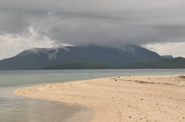 Sakin bir sahil sahnesi, sakin bir denize giden yumuşak kumu yakalar. Kara bulutlar uzak bir dağın üzerinde süzülürken, muhtemel yağmuru işaret eder..