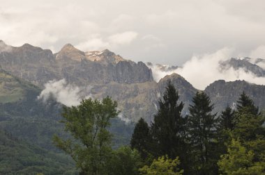 Zirvelerin etrafında bulutların dolaştığı engebeli dağ zirveleri hakim. Ön planda duran uzun ağaçlar bulutlu bir günde vadinin sakin atmosferini vurguluyor..
