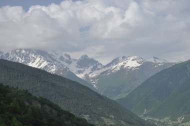 Nefes kesici bir manzara bulutlu bir gökyüzünün altında kar parçalarıyla süslenmiş yüksek bir dağ sırasına sahiptir. Aşağıdaki yemyeşil vadi, doğanın güzelliğini gözler önüne seriyor..