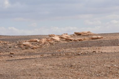 Dağılmış açık renkli kaya oluşumlarına sahip bir çöl arazisi. Gökyüzü parçalı bulutlu, kurak arazide dramatik bir zemin oluşturuyor..