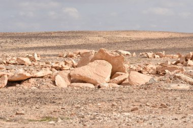 Kurak bir arazi, açık mavi bir gökyüzünün altında, yeryüzündeki erozyon şekillerini ortaya çıkaran, dağılmış kaya ve taşları barındırır. Kuru, kayalık zemin arka planda durmaksızın uzanıyor..