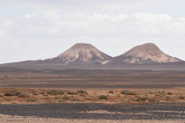 İkiz dağlar, kuru toprak ve seyrek bitki örtüsüyle çevrili uçsuz bucaksız bir çöl arazisinde göze çarpıyor. Yukarıdaki bulutlar sert manzaraya derinlik katıyor..