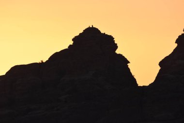Yürüyüşçüler gün batımında bir dağın zirvesinde, gökyüzünde nefes kesen renklerle çevrili dururlar. Engebeli manzara, sıcak parıltıya karşı çarpıcı siluetler oluşturur..