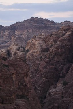 Petra yakınlarındaki sönen ışıkta engebeli dağ oluşumları dimdik ayakta duruyor. Alacakaranlık, kahverengi ve gri tonlarını ortaya çıkarır. Arazinin doğal güzelliğini ve eşsiz dokusunu vurgular..