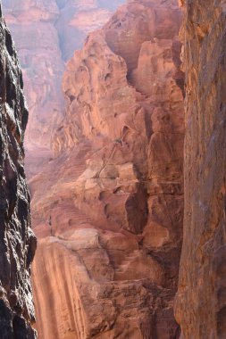 Nefes kesici bir manzara, açık mavi gökyüzünün altında dar bir kanyonu çerçeveleyen dik kırmızı kaya duvarlarını gözler önüne seriyor. Güneş ışığı canlı renkleri aydınlatır, doğanın güzelliğini detaylarıyla gösterir..