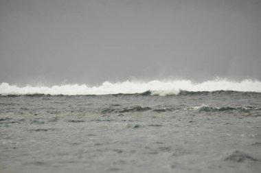 Büyük dalgalar tenha bir sahile yuvarlanırken kara bulutlar uzanıyor ve kıyıya vuran suyun ritmik sesiyle sakin bir atmosfer yaratıyor..