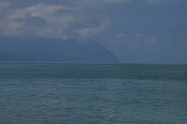 Sakin okyanus suları ufka kadar uzanıyor, mavinin tonlarını yansıtıyor. Uzak dağların üzerinde kara bulutlar beliriyor, sakin ama dramatik bir atmosfer yaratıyor. Sahne doğanın güzelliğini yakalar..