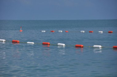 Parlak turuncu ve beyaz şamandıralar suda yüzerek yüzücüler için görünür bir bariyer oluştururlar. Sakin deniz, berrak bir gökyüzünü yansıtır ve gün boyunca huzurlu bir ortam yaratır..