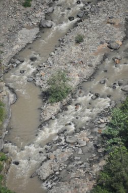 Huzurlu bir nehir yeşilliklerle çevrili kayalık bir arazide kıvrılır. Su biraz çamurlu görünüyor. Bu da yakın zamanda yağdığını ya da yakınlarda olduğunu gösteriyor..