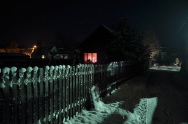 Geceleri huzurlu bir mahallede kar ve çitler kaplanır. Bir evin penceresinden parlayan ılık bir parıltı, soğuğun ortasında rahat bir atmosfer yaratır..