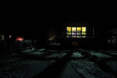 Sıcak bir ev, kış gecesinin karanlık zeminine karşı renkli ışıklarla parıldıyor. Kar zemini kaplıyor, huzurlu ve huzurlu bir atmosfer yaratıyor..