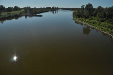 Parlak mavi bir gökyüzünün altında huzurlu bir nehir akmaktadır. Güneş yemyeşil ve uzak ağaçlarla çevrili sulara yansıyarak huzurlu bir manzara yaratıyor..