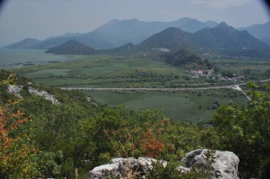 Parlak yeşil tepeler ön planda uzanır, berrak mavi gökyüzünün altındaki dolambaçlı bir nehir vadisine uzanır. Dağlar uzaklardan yükselir ve huzurlu bir açık hava manzarası yaratır..
