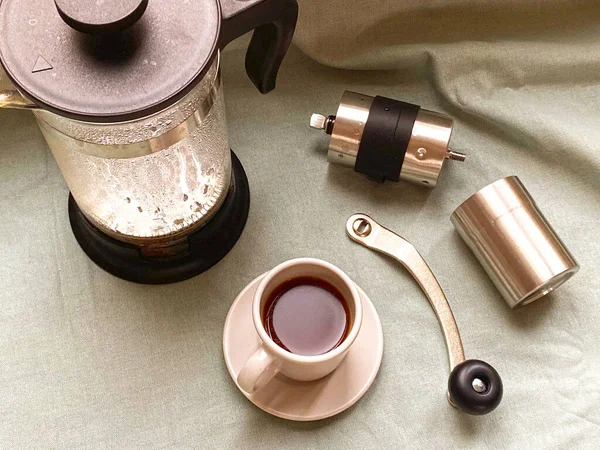 Vaporous wet french press, coffee cup with dark hot espresso on saucer and small metal coffee grinder on light green fabric cloth. Still life. Making of fresh coffee at home.