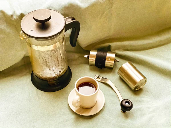 Vaporous wet french press, coffee cup with dark hot espresso on saucer and small metal coffee grinder on light green fabric cloth. Still life. Making of fresh coffee at home.