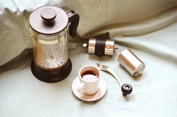 Vaporous wet french press, coffee cup with dark hot espresso on saucer and small metal coffee grinder on light green fabric cloth. Still life. Making of fresh coffee at home.