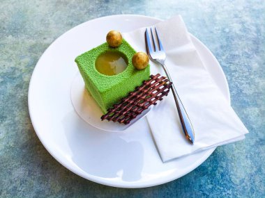 Üzerinde altın küreler olan yeşil armut küplü pasta ve yanında mavi bir masada beyaz bir tabakta çikolata ızgarası.