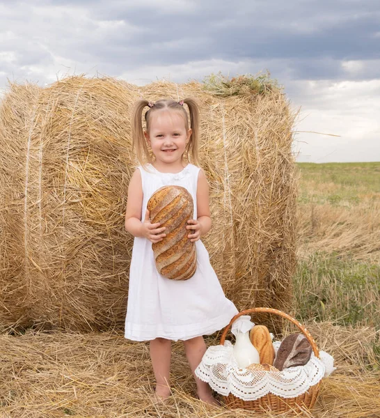 Маленькая блондинка в белом платье держит хлеб в поле. экологически чистая сельскохозяйственная продукция. — стоковое фото