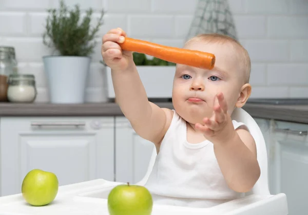 Healthy baby boy sitting in a childs chair in the kitchen eats orange carrot. — Stock Photo, Image