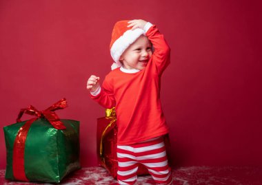 Kırmızı Noel Baba şapkalı çocuk. Kırmızı arka planda Noel hediyeleri var..