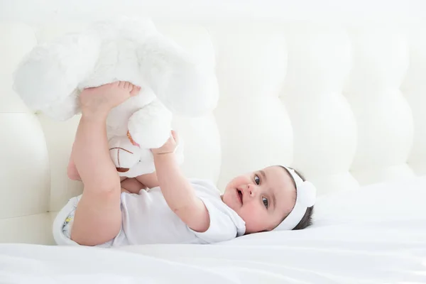 Divertida niña de 6 meses jugando con las piernas, acostada en la cama en casa sobre ropa de cama blanca. vista superior. — Foto de Stock