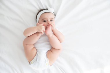 Komik 6 aylık bebek bacağıyla oynuyor, beyaz yatakta yatıyor. en üstteki görünüm.