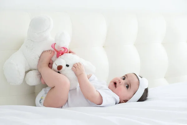 Healthy baby girl 6 months in a white bodysuit lying on a bed on white bedding with teddy bear. — Stock Photo, Image