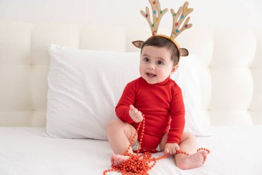 6 aylık kırmızı bir vücut giysisi ve boynuzları beyaz bir yatakta oturan bir geyik boynuzuyla..