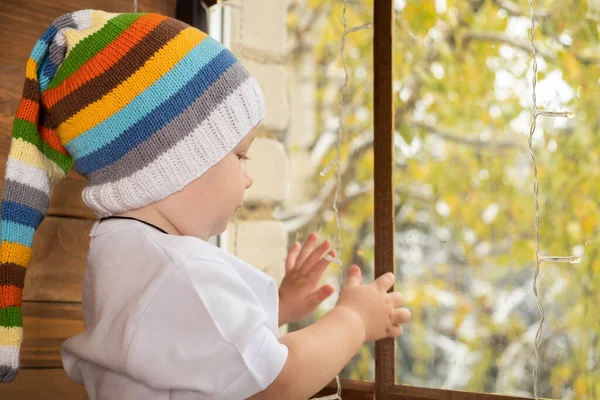 Mały chłopiec w kolorowym kapeluszu patrzy przez okno. — Zdjęcie stockowe