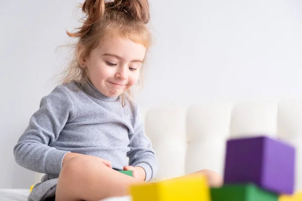 Маленькая девочка играет с красочными деревянными кубиками, сидя на кровати. Домашние мероприятия для детей. — стоковое фото