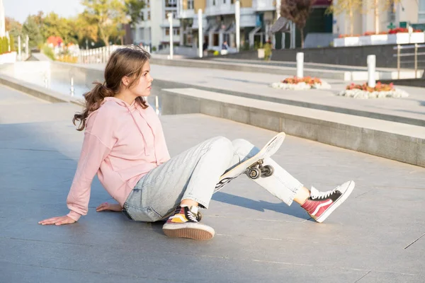 Mladý alternativní teenager dívka bruslař sedí na palubě na ulici města dívá pryč. — Stock fotografie