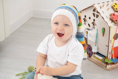 Komik şapkalı, beyaz tişörtlü, evde oyuncak ayıyla oynayan bebek..