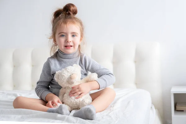 Menina em gola alta cinza com ursinho de pelúcia sentado na cama sorrindo. — Fotografia de Stock