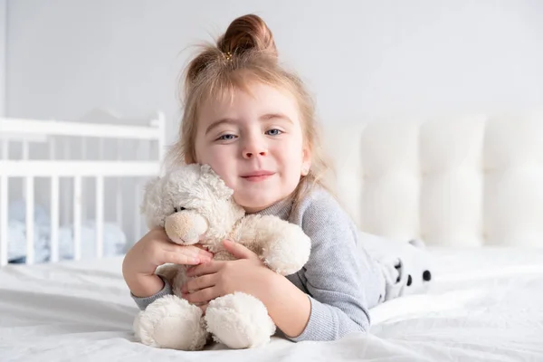 Menina em gola alta cinza com ursinho de pelúcia que coloca em roupa de cama branca sorrindo. — Fotografia de Stock