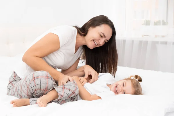Volwassen moeder kietelt haar kleine actieve dochter in bed thuis, vrijetijdsbesteding met kinderen. — Stockfoto