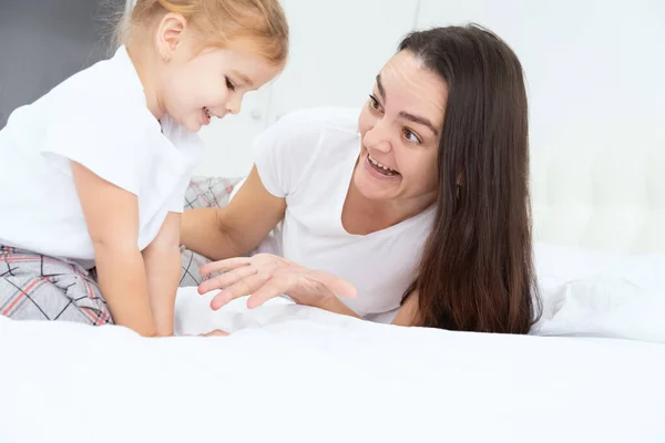 Volwassen moeder spelen met weinig actieve dochter in bed thuis, plezier hebben, activiteit met kinderen. — Stockfoto