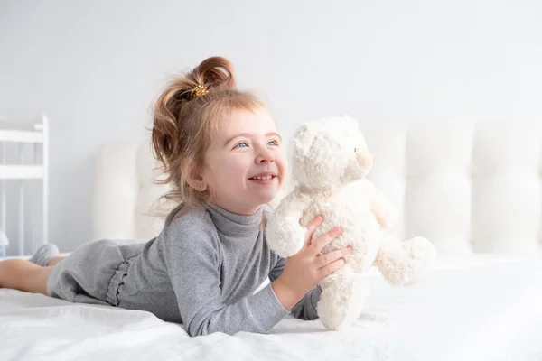 Menina em gola alta cinza com ursinho de pelúcia que coloca em roupa de cama branca sorrindo. — Fotografia de Stock