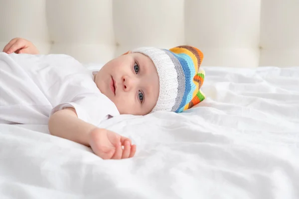Menino engraçado em chapéu colorido engraçado sorrindo e deitado em uma cama branca em casa. — Fotografia de Stock