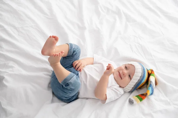 Funny baby boy in funny colorful hat smiling and lying on a white bedding at home. — Stock Photo, Image