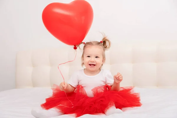 Mignon bébé fille tient ballon de coeur est assis sur une literie blanche. — Photo