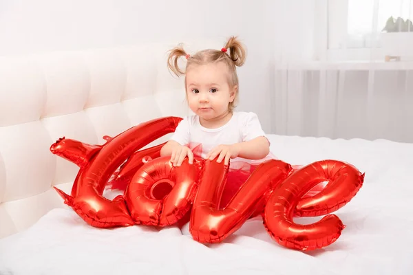Roztomilá holčička drží nápis láska z balónky sedí na bílém lůžkoviny. — Stock fotografie