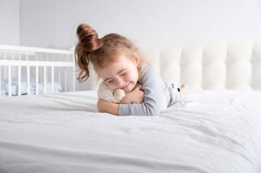 Gri balıkçı yakalı küçük kız beyaz yatakta oyuncak ayıyla oynuyor.