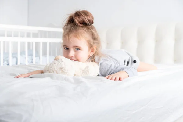 Menina em gola alta cinza brincando com ursinho de pelúcia na cama branca — Fotografia de Stock