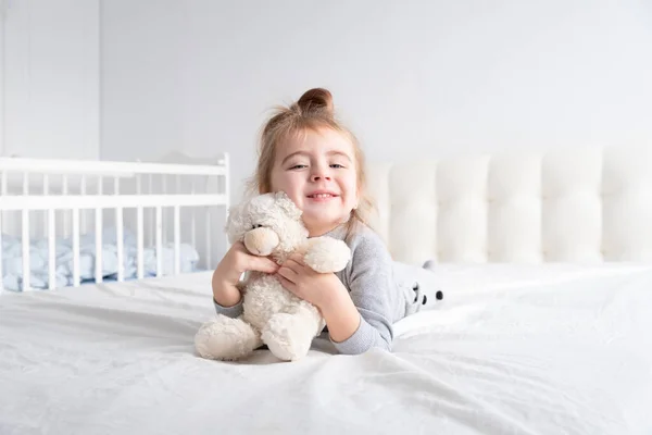 Menina em gola alta cinza brincando com ursinho de pelúcia na cama branca — Fotografia de Stock