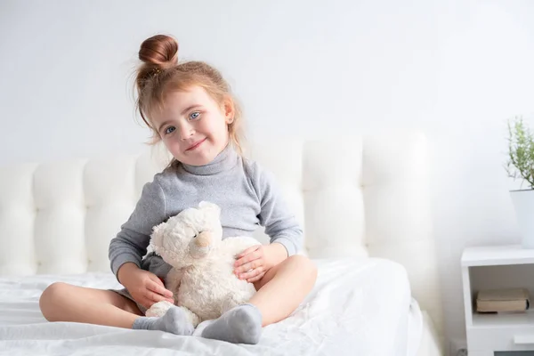 Menina em gola alta cinza brincando com ursinho de pelúcia na cama branca — Fotografia de Stock