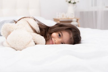 Küçük esmer kız evde oyuncak ayıyla yatakta yatıyor..