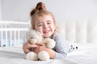 Gri balıkçı yakalı küçük kız beyaz yatakta oyuncak ayıyla oynuyor..