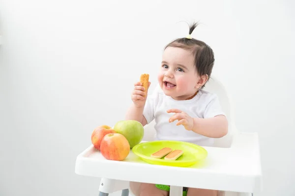 Bebek koltuğunda oturan bebek beyaz arka planda elma ve kurabiye yiyor. Bebek önce katı yiyecekler