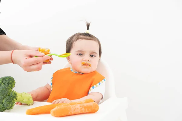 Childs sandalyesinde oturan küçük kız beyaz arka planda sebze püresi yiyor. Anne bebeği besliyor.